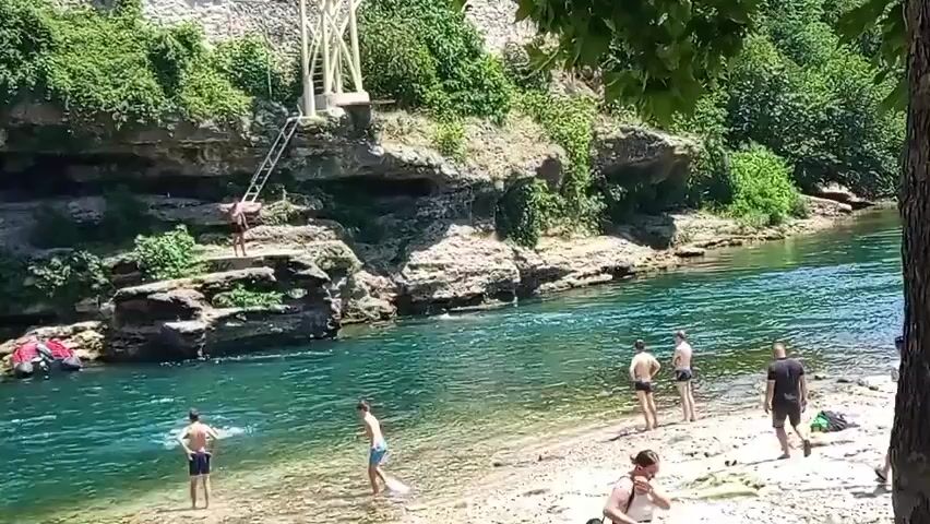 A wide shot shows a clear, turquoise river flowing through a rocky, tree-lined gorge on a sunny day. Several people are visible on the sandy bank to the right, some standing, others walking, and one sitting. Two individuals are wading in the shallow water near the left bank. In the background, on the left side of the river, a person is climbing a ladder attached to a rock face that has a white, elevated structure at its summit. Another person is on a ledge on the rock face, preparing to jump into the water.