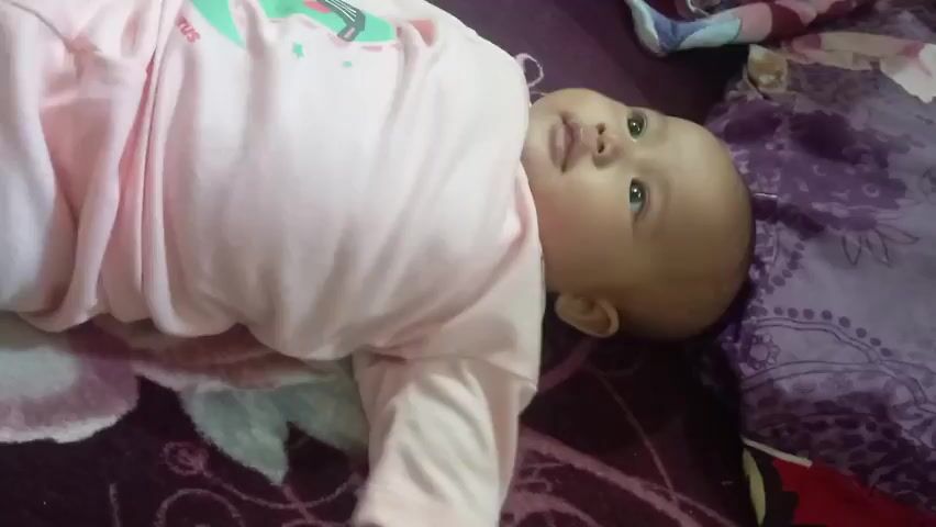A baby is lying on its back on a bed. The baby is wearing a light pink onesie with a green dinosaur graphic on the front. The baby's head is turned to the right, and its eyes are open, looking upwards. Its left arm is bent at the elbow, and its hand is resting on its chest. Its right arm is extended downwards. A person's feet, with red nail polish, are visible in the lower left corner of the frame. The bed is covered with a dark purple patterned blanket. A large, bright pink object, possibly a toy or blanket, partially obscures the baby from the top.