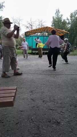 A group of older adults are dancing outdoors near a small, teal-colored stage with a yellow awning. A man dressed in a dark uniform and hat stands on the stage, appearing to lead the dance. A woman in a bright yellow top and brown pants is dancing enthusiastically in front of the stage. Other individuals are scattered around, some dancing and some observing. One person in the foreground is holding up a phone, seemingly recording the event. The ground is paved with asphalt and gravel. Trees and foliage are visible in the background.