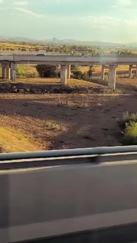 The video begins with a view of a dry, barren landscape with a bridge overhead. Cars can be seen driving on the bridge. The camera then shifts to the interior of a vehicle, showing a person's head and shoulders as they look out a window. The window reflects the interior of the vehicle, with some lighting elements visible. Outside the window, the sky is clear and blue. An airplane is visible flying in the distance. There are also some buildings and a cell tower visible in the background. A person is seen holding a smartphone and taking a picture of the airplane. There are some stickers on the window. The video ends with a close-up of the person's hands holding the phone.