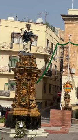The video pans across a street scene, possibly in a European city, with a focus on festive decorations. A statue of a saint stands prominently on an ornate pedestal in the foreground. The statue is white with a black garment and a halo. The pedestal is gilded and elaborately decorated. Green garlands are strung across the street, adorning buildings and lampposts, some of which have small lights. The buildings are made of stone and have traditional architecture with balconies and windows. There are also flags visible on some buildings. In the background, there are glimpses of other statues, street furniture like umbrellas, and some people walking or standing. The overall impression is a celebratory atmosphere, perhaps for a festival or religious event. The lighting suggests it is daytime with clear skies.
