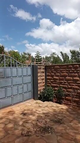 The video shows a low brick wall and a metal gate in front of a property. The wall is made of tan-colored bricks, with dark gray columns on either side of the gate. A black lamppost sits atop one of the columns. A tall, thin evergreen tree grows behind the wall, along with other leafy trees and glimpses of a red-tiled roof. The ground in front of the wall is covered in irregular stone paving. The sky is blue with scattered white clouds. The camera zooms slightly to focus on the evergreen tree.