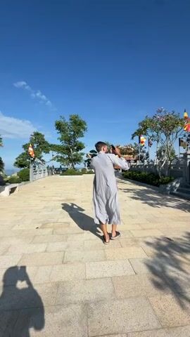 The video shows a person in a gray robe walking towards a golden tower on a sunny day. The tower is multi-tiered and has red flags flying next to it. Then, the video changes perspective to show another person walking on a paved surface, casting a shadow. Finally, the video shows a woman in a gray robe taking a selfie, smiling at the camera. In the background, there are trees, an umbrella, and a bench.