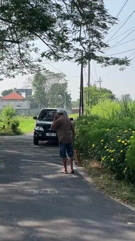 A man in a tan shirt, denim shorts, a white cap, and flip-flops walks along the right edge of an asphalt road, facing away from the camera. He holds a broom in his right hand and appears to be gesturing with his left hand towards his head. To his left, a black SUV is parked on the side of the road. The SUV has a license plate that is partially visible. The road is flanked by lush green vegetation on both sides. In the background, there are trees, power lines, a utility pole, and a building. The sky is overcast.