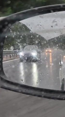 This media file captures a view from a car driving on a highway in Gorno Kamartsi, Bulgaria. The scene is dominated by a rainy and overcast day, with water droplets visible on the side-view mirror in the foreground. The mirror reflects traffic moving on the wet highway, including several cars with their headlights on and the illuminated reflections of their lights on the road surface. In the background, lush green hillsides are visible, suggesting a rural or mountainous environment. The highway itself appears to be a multi-lane road with a guardrail separating lanes. A dark-colored car is prominently featured in the right lane, its wheels splashing water as it moves. Further ahead, a large blue shipping container is being transported by a truck. The overall mood conveyed is one of driving through adverse weather conditions.