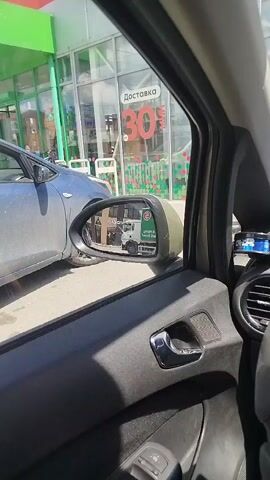 The video, filmed from inside a car, shows a view through the passenger window. In the foreground, the car's interior is visible, including a side mirror, door handle, and part of the steering wheel. The side mirror reflects a white truck. Outside the car, a grey car is parked to the left. Behind it is a building with large glass windows displaying advertisements. One prominent advertisement reads "ДОСТАВКА 30%". A man wearing a white cap and a dark t-shirt, pushing a shopping cart, is visible through the glass. He is standing near the entrance of the building. The environment appears to be a commercial area on a sunny day.