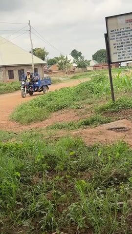 A tricycle with two passengers drives down a dirt road in an outdoor environment. The road is flanked by green vegetation and some buildings. A sign is visible on the right side of the road. The sky is overcast. The tricycle then passes a building under construction.