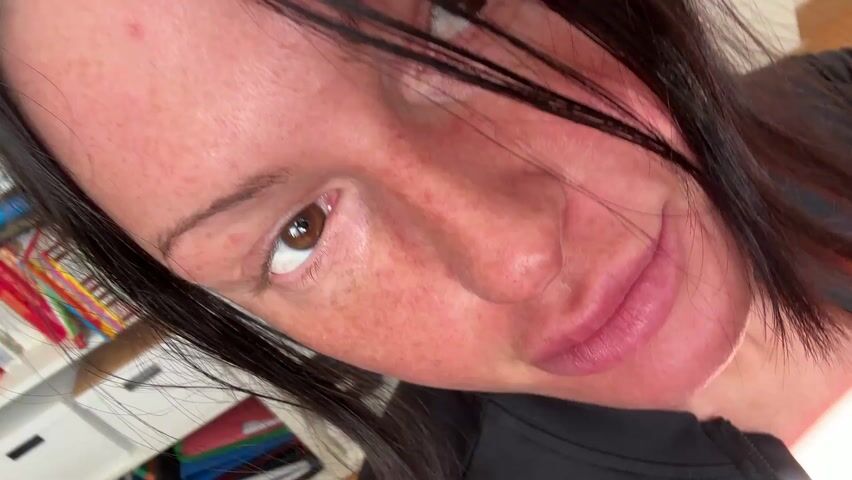 A woman with brown hair and freckles is the primary subject of this video. She's wearing a pink athletic top and black workout gear, including leggings and a long-sleeved jacket. The camera seems to be held at a low angle, focusing on her face as she looks directly at the lens. In the background, there is a white shelving unit and sunlight filtering through a window, creating shadows in the room. The camera angle and movement suggest a casual, self-shot video.