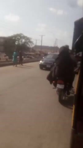 The video shows a street scene with traffic. A black Mercedes-Benz station wagon is parked on the left side of the frame. Several motorcycles are seen driving on the road, some with passengers. A red car drives by in the distance. A yellow tricycle is visible on the right side of the frame, from which the video seems to be recorded. The sky is overcast and there are buildings and trees in the background. The overall environment suggests a busy urban street.