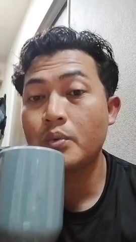 A person takes a sip from a gray mug. The person is wearing a black t-shirt. The background is a plain wall.