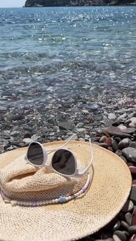 A close-up shot of a straw hat and sunglasses lying on a pebble beach. The water is clear and shallow, with waves gently lapping at the shore. In the background, a green, tree-covered hill rises from the sea under a clear blue sky.