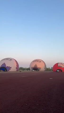 The video shows hot air balloons partially inflated on a maroon-colored field under a clear blue sky. The balloons are branded with "volare.com.mx" and "XB-BDQ" in addition to some other branding. There are people and cars near the balloons. One partially inflated balloon is also seen floating in the distance.