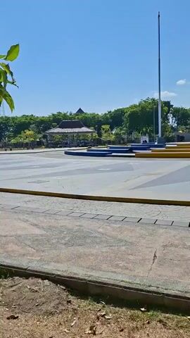 The video shows a bright, sunny outdoor scene with a clear blue sky. The foreground features a paved area with square paving stones and a dirt and grass border. To the right, a dark metal bench with decorative cutouts is visible. In the midground, there is a large, open, paved plaza with geometric designs. Several low, blue, rectangular platforms are arranged on the plaza. A tall flagpole stands to the left, and further back, two tall, cream-colored obelisk-like structures rise above a stage or elevated platform. Behind these structures, there is lush green foliage and trees. A covered pavilion with a traditional thatched roof is visible among the trees on the left side of the midground. The words "ALUN ALUN" are spelled out in large blue letters on a raised platform in the background. The overall impression is that of a public park or town square on a clear day.