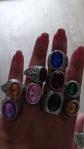 A person's hand, adorned with ten colorful rings, is displayed against a blurred background. The rings feature large, faceted gemstones in various colors, including blue, red, green, pink, purple, and orange. The hand, with manicured fingernails, is positioned in a way that showcases the collection of rings. The environment appears to be indoors, with a hint of a wooden surface in the background.