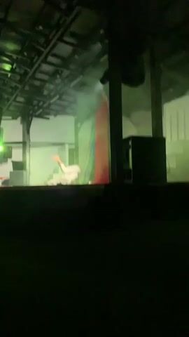 A performance is taking place on an outdoor stage under a covered roof. The stage is lit with dramatic colored lights, primarily red and purple, with some smoke effects. There are two individuals on the stage, dressed in light-colored clothing. They appear to be dancers or actors, engaged in choreographed movements. One individual is on the left side of the stage, partially seated. The other individual is on the right, also in a dynamic pose. In the background, there are white structures, stairs, and a red and blue curtain. A sign that reads "TEATRO PALMERAS" is visible on the left side of the stage. The audience is not visible, as the foreground is dark.