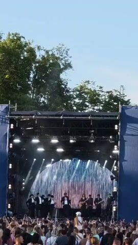 A group of musicians perform on a stage with a sparkly, curtain-like backdrop. The stage is lit with bright lights, and a scaffolding structure surrounds it. In front of the stage, a large crowd of people are gathered, many holding up their phones. Trees and a clear blue sky are visible in the background.