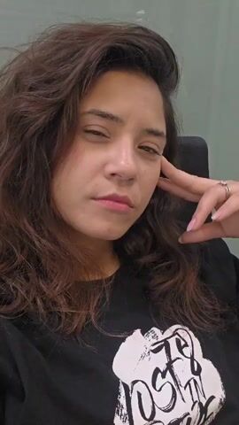 A person, with brown wavy hair and light brown skin, sits in a black office chair, wearing a black T-shirt with white graphics. They are looking directly at the camera with a slight smile. They close their eyes briefly, then smile again. They then make a kissy face and give a peace sign with their right hand. The background appears to be a plain office wall, possibly green.