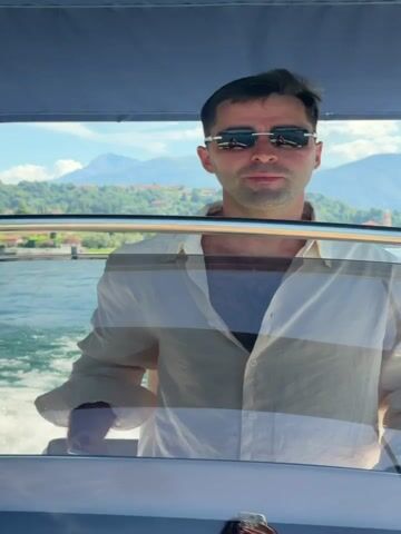 The video shows a man in sunglasses operating a boat. He is wearing a light-colored, open-collared shirt over a darker garment. The boat appears to be moving on a body of water, with waves visible. In the background, there are mountains and trees. The man looks around and occasionally down at a brochure or paper he is holding. The lighting suggests it is daytime.