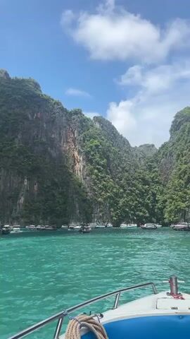 The video shows a first-person view from the front of a white motorboat moving through clear turquoise water. Towering, lush green cliffs rise steeply on either side, forming a narrow passage. Numerous boats, including speedboats and smaller vessels, are anchored or moving within the cove. The sky is blue with scattered white clouds. The water is calm with gentle ripples. The boat's railing and a coiled rope are visible in the foreground.