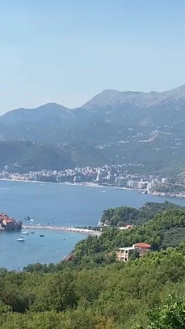 The video pans across a coastal landscape under a clear blue sky. In the foreground, lush green trees form a dense canopy. Beyond the trees, a wide expanse of blue water, the sea, stretches out. In the middle distance, a small island with red-roofed buildings and a bridge connecting it to the mainland is visible. Several boats, including a white yacht, are scattered on the water around the island. Further in the background, a coastal city with numerous buildings and a mountainous backdrop can be seen. The overall scene depicts a sunny day at a scenic coastal resort.