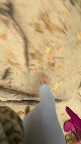 A person with bare feet walks on a sandy surface covered with tree roots and scattered leaves. The person wears light blue pants and a camouflage-patterned bag. A pink object is visible in the corner. The camera follows the person's feet while they walk.