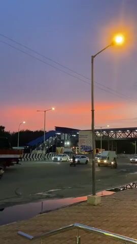 This is a vertical, eye-level shot of a street scene at dusk. The sky is a gradient of deep blue at the top, transitioning to a vibrant orange and pink near the horizon, indicating sunset. A single streetlamp on the right side of the frame is illuminated, casting a warm glow.

The foreground shows a paved walkway with a textured surface, bordered by a curb. Beyond that is a wet asphalt road, reflecting the sky and streetlights. A small puddle of water is visible on the road.

In the middle ground, a busy intersection is captured. Cars, a white truck, and several motorcycles are navigating the road. A pedestrian overpass, painted blue and grey, spans the road in the background, with people visible on it. The overpass is illuminated by lights, creating a contrast with the darkening sky. Road dividers with black and white markings are prominent.

To the right of the overpass, a white sign with text and graphics is mounted on a pole. The text appears to be in Tamil, with some English words like "Bus Service" and "Timings" discernible. This suggests the location is a public transport hub, possibly a bus station.

The overall mood is serene and tranquil, enhanced by the beautiful sunset colors and the soft glow of the streetlights. The presence of vehicles and pedestrians indicates it's an active urban environment, even as daylight fades. The location context places this scene in St. Thomas Mount-Pallavaram Cantonment Board, India.