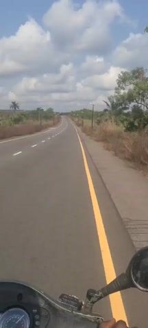 The video shows a first-person view of a vehicle traveling down a paved road. The road is straight with a slight incline.  There is a white line dividing the two lanes and a yellow line along the right edge of the road. Streetlights line the road. Vegetation borders both sides of the road, including palm trees. The sky is cloudy with patches of blue.