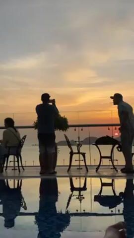 The video shows a group of people silhouetted against a sunset. There are three distinct individuals discernible. One woman is seated at a table and two men are standing by a glass-paneled balcony railing. One man is holding up a phone and photographing the sunset. The other man is standing and looking out at the sunset, with a cigarette in his mouth. There is a table and several chairs near the railing. The reflection of these figures and furniture can be seen in the pool below. The sky is painted with warm tones of orange and yellow as the sun appears to be setting over the water. Islands are faintly visible in the distance.