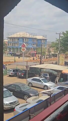 Here is a description of the video:

The video shows a view from inside a building, looking out onto a busy street scene in what appears to be a city in Nigeria. There are multiple cars parked in a lot below, along with a covered area. In the distance, a prominent blue and white building stands out, featuring advertisements for Tecno and Chicken Republic. People and motorcycles are moving along the street. The camera then shifts to show electrical wires and part of the building's interior. The video then pans across the street, revealing more parked vehicles, motorcycles, and people. Street vendors with stalls can also be seen. The overall impression is of a bustling urban environment.