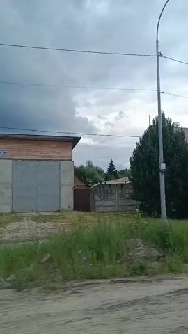 The video pans across a rural landscape under a cloudy sky. In the foreground, a dirt road is visible. On the left, a large building with a concrete base and a wooden upper section and a large metal door appears to be a garage or industrial building. To the right of this building is a house with a red roof. Further down the road, there is a long, dark-colored fence with a sign that reads "СТРОЙМАТЕРИАЛЫ" (which translates to "Building Materials"). Beyond the fence, trees and vegetation are abundant. The camera continues to pan, revealing a small, gray house with a white trim, situated in a grassy area. A pile of sand or gravel is also visible. The video ends with a view of more trees and greenery, indicating a heavily wooded area. The overall scene suggests a village or rural industrial area.