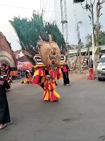 The video shows a street performance with people in elaborate costumes. The costumes appear to be large and animal-like, with furry heads, exaggerated features, and colorful, striped fabrics. Some of the performers are wearing large headdresses with feathers and decorative elements. There are also people in the background, some watching the performance and others moving around the area. Vehicles, including scooters and cars, are visible on the street, suggesting an outdoor public event. The overall atmosphere appears to be festive and lively.