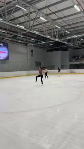 The video shows three ice skaters on an indoor rink. The skater in the foreground is wearing a purple long-sleeved shirt and black pants. She performs spins with her arms extended and then raises them overhead. Two other skaters are visible in the background. The rink is surrounded by a low yellow barrier, and a large screen displays text and images. The building has a high ceiling with visible metal support beams. The ice is smooth and shows some skate marks.