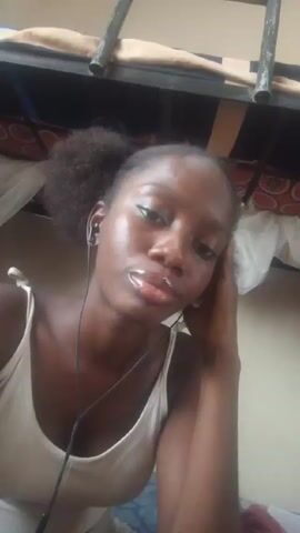A close-up selfie video shows a young, dark-skinned woman with her hair pulled back into a ponytail, wearing a white tank top. She is looking at the camera and smiling slightly. She is wearing in-ear headphones. The background shows the underside of a bunk bed. There is background noise, including a voice speaking.
