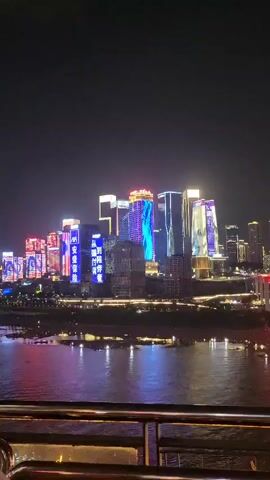 The video shows a nighttime cityscape with numerous skyscrapers illuminated by bright, colorful lights. The buildings are of varying heights and designs, some featuring vertical LED displays showing patterns and text. In the foreground, a railing with metal bars is visible, suggesting the viewpoint is from a balcony, deck, or bridge. Below the cityscape, a body of water reflects the vibrant lights, creating a colorful, shimmering effect. The water appears calm, with subtle ripples. The sky is dark, emphasizing the illuminated buildings and their reflections. The overall scene is one of a modern, bustling city at night, characterized by its vibrant urban lighting.