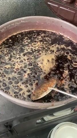 A close-up, high-angle shot shows a large, round, metal pot on a stovetop, filled with dark purple-black berries or fruit simmering in a viscous liquid. The liquid is actively bubbling, creating a light brown foam on the surface. A metal slotted spoon with a brown, textured head is inserted into the pot and is actively stirring the fruit mixture, moving it from the center towards the right side of the pot. In the upper right background, a brown, tiered rack holding several small, dark jars or containers is visible. A white plastic bucket or container is partially visible in the bottom right foreground.