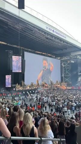 A wide shot shows a concert stage with a massive screen displaying a performer. The performer is a man in a black outfit with a microphone in his hand. He is the central focus of the screen. The stage itself has decorative elements. In front of the stage, a large crowd of people are gathered, many holding up their phones to record. The concert appears to be held in an open-air stadium, as indicated by the presence of stadium seating and a large roof structure overhead. The sky is visible above the stadium. The overall atmosphere suggests a large-scale musical performance.