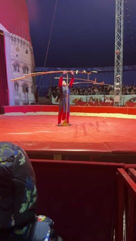 A circus performer stands on a red platform, balancing long sticks above his head. He is dressed in a dark, long-sleeved tunic over red pants. The background includes an audience seated in rows and a painted backdrop of a building. A tall metal structure is visible to the right, possibly part of the circus rigging. A spectator sits in the foreground, their back to the camera.