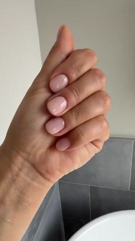 The media file contains a close-up shot of a person's hand with light pink, well-maintained nails. The hand is positioned in various poses against a backdrop of what appears to be a bathroom. The background shows grey tiled walls and a white sink, suggesting an indoor setting, likely a bathroom in Houston, Texas, given the location context. The lighting is soft and diffused, indicating it could be daytime, but the specific time is not discernible. There is no visible text in the media file. The focus is entirely on the hand and its nails, which are neatly shaped and polished, implying care and attention to grooming. The overall impression is one of neatness and a simple, elegant aesthetic.