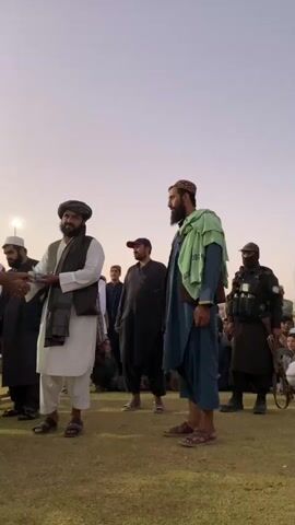 The video begins by showing a young male soccer player in a green and white uniform shaking hands with an adult male wearing a white shalwar kameez, a dark vest, and a turban. The player is holding a trophy. Behind them, other people are gathered, some are seated and some are standing. In the background, there are more trophies on a table. The scene appears to be an awards ceremony taking place outdoors on a grassy field. The sky is a light orange and pink, suggesting it is either sunrise or sunset. As the video progresses, the player receives the trophy from the man. The man then shakes hands with another adult male who is wearing a blue shalwar kameez and a green sash over his shoulder. To the far right, a man wearing tactical gear and holding a rifle is visible. The camera is positioned at a medium distance, capturing the interactions and the surrounding environment.