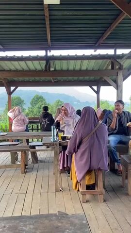 People are seated at picnic tables in a covered, outdoor structure made of wood.  Several people are wearing hijabs.  Some are eating or drinking. The structure has a wooden deck floor and a green metal roof. Beyond the deck is a view of distant mountains.