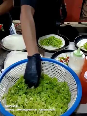 Here is a detailed description of the video.

A man wearing black gloves is preparing food at a food stall. He's placing shredded lettuce from a blue basket onto round wrappers on black plates. There are four plates in total, each with a wrapper and some lettuce. One plate also has some red sauce. Next to the plates, there are squeeze bottles containing sauces. The food stall is visible in the background, with another person working on the left side. The video has text overlayed at the bottom indicating it was taken with an OPPO A6 Pro.