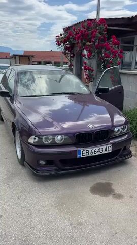 The video shows two BMW cars parked on an asphalt street in a village. The first car, closer to the camera, is a dark purple sedan with Bulgarian license plates. The front passenger door is open.  Behind it is a white hatchback, also with Bulgarian license plates.  A residential building with a red tile roof and a rose bush is visible behind the purple car.  The sky is blue with scattered clouds, and distant hills are visible in the background. The camera pans slightly to follow the cars.