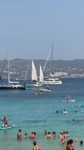 A wide shot captures a sunny day at a beach with clear, turquoise water. Several sailboats and a catamaran are anchored in the bay. In the foreground, numerous people are enjoying the water, with some swimming and others on paddleboards. One man is paddling a green paddleboard, while another is standing on a similar board further out. In the background, a hillside with buildings and trees is visible under a clear blue sky.