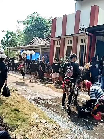 The video shows a group of people gathered outdoors in what appears to be a village or rural area. A building with a red and white facade is visible in the background, along with trees and vegetation. Several individuals are dressed in dark clothing, some wearing black shirts with logos or symbols. One person in the foreground is wearing a white cap and a black outfit, observing the scene. Another person on the right is dressed in traditional attire, possibly for a performance or ceremony, and is interacting with others. In the center of the scene, water is being thrown, creating splashes and wetting the ground. Buckets are also visible, suggesting water may be used for a ritual or cleansing activity. The overall atmosphere seems to be that of a gathering or event, possibly with cultural or traditional elements.