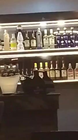 The video begins focused on two shelves of liquor bottles illuminated by strip lighting. An ice bucket is visible on the bar in front of the shelves. The camera moves to a dark bar surface and then quickly pans to a blue decorative light fixture.
