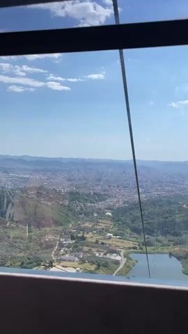 The video shows a panoramic view of a city and its surrounding landscape from a high vantage point, likely from a cable car or observation deck. The sky is blue with scattered white clouds. In the foreground, the edge of a dark-colored structure, possibly a window frame or cable car interior, is visible. A thin black cable runs vertically through the center of the frame, suggesting the viewpoint is from a cable car. Below, a sprawling city with numerous buildings stretches into the distance, meeting rolling hills and mountains on the horizon. Lush green vegetation covers the lower slopes of the hills, with some scattered houses and structures visible. A body of water, possibly a lake or reservoir, is present in the mid-ground, with a pathway or road running alongside it. The scene is illuminated by natural daylight, indicating it is daytime. There are no people or animals clearly visible in the frame. The overall impression is one of an expansive urban and natural landscape.
