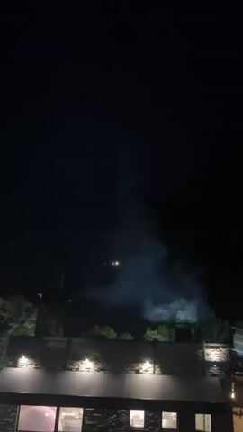 The video shows a night scene with smoke rising into the air. In the distance, there are several lights of different colors (red, orange, green, purple) moving around. In the foreground, there is a low stone wall with lights on it, and some greenery. The sky is dark.