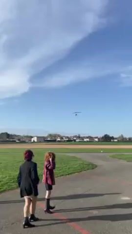 Three people stand on an asphalt path in front of a grassy field, watching a glider descend through the sky. The glider is a dark color and flies toward the left side of the frame. One person stands on the path holding a controller of some kind, presumably to control the glider. They wear a black jacket, gray pants, and a baseball cap. Two other people, dressed in fashion-forward clothing, stand next to them. The person on the left has short dark hair and wears a black blazer, dress, and black booties. The person on the right has curly red hair and wears a red jacket, black shorts, and black boots. Behind the field, there is a cluster of houses and trees. The sky is blue with some clouds. The lighting is bright and sunny.
