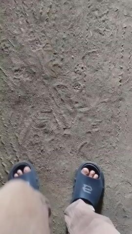 The video shows a first-person perspective of someone walking on a dry, dusty ground. The person is wearing light-colored pants and dark blue slides. The camera is focused on the person's feet as they walk. The ground appears to be a mix of dirt and small pebbles, with some dry leaves scattered around. The lighting suggests it is daytime. The camera movement is slightly unsteady, indicating a natural walking motion.