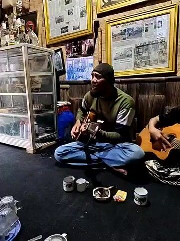 The video shows two musicians performing in a room filled with various objects.

One man with a beard and wearing a dark beanie, a green and white long-sleeved shirt, and blue jeans sits cross-legged on a black rug, playing an acoustic guitar. Beside him, another musician plays a second guitar. In front of them on the rug are several white mugs, an ashtray, and a small box of matches.

The camera then shifts focus to a third man with long dark hair tied back. He is wearing a green t-shirt and dark pants, kneeling on the floor and playing a small hand drum.

The room is cluttered. Behind the musicians, two framed newspaper articles hang on a wall covered in bamboo matting. A glass cabinet displays various items on shelves. The room also contains other objects, including a fish tank, a blue drum, and a variety of decorative and household items. The lighting is soft and warm, creating a cozy atmosphere.
